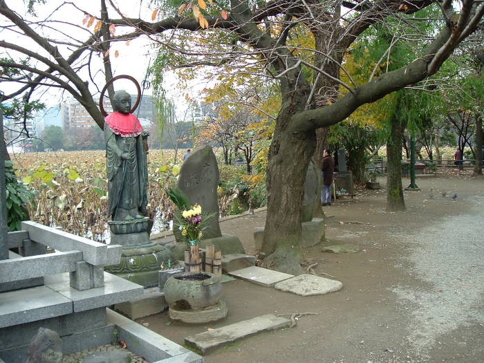 Red-bibbed statue of the Buddhist deity Jizō at Benzaiten Buddhist Temple. The deity is depicted as a monk with a staff in one hand and a jewel in the other. Someone has placed a red bib on the figure.