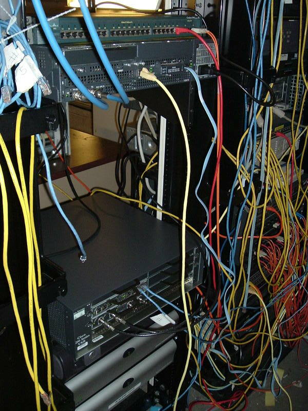 Yellow and orange network cables and fiber optic lines hanging from stacks of networking equipment in multiple racks.