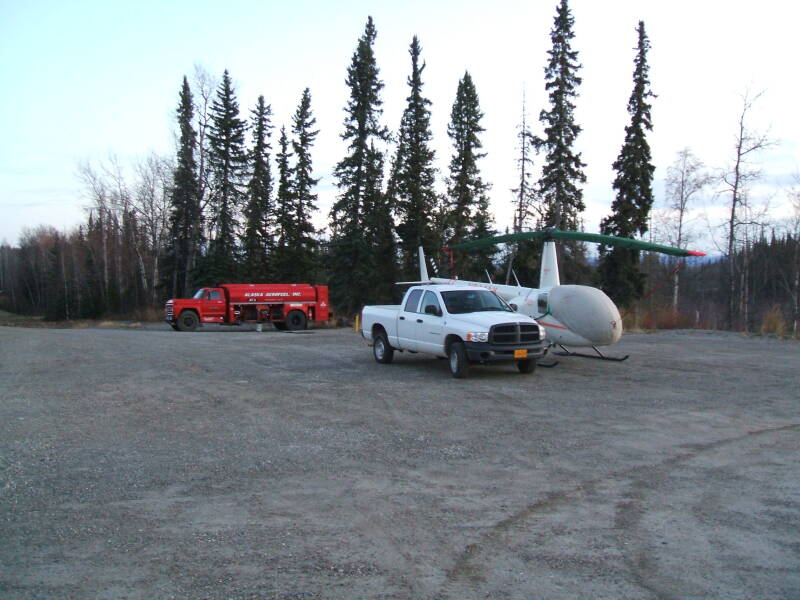 Helicopter parking in Alaska.  A helicopter, a fire truck, a pickup truck, and pine trees.