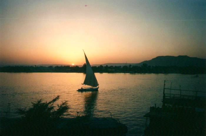 Sunset over the Nile River and a falucca at Luxor, the Valley of the Kings is in the distance at right.