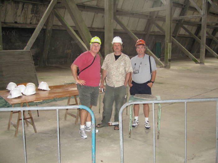 Three visitors in hard hats, inside a concrete balloon hanger.