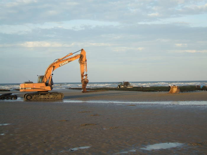 Excavator working on Utah Beach, moving steel oyster frames near the low-water line.