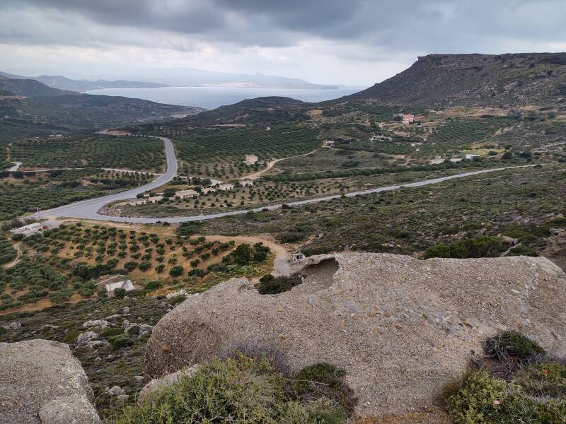 A twisting narrow road descends a mountain, joining the main road which leads on to Sitia, seen in sunlight in the distance.