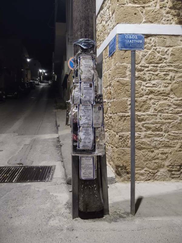 Utility pole with several funeral notices, at night along a street in Sitia.