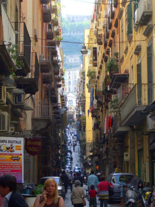 A narrow street in Napoli's Centro Storico slopes steeply uphill between buildings with apartments upstairs and shops at street level.