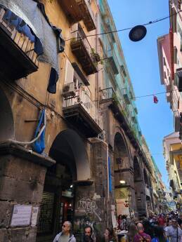 Many people walking through the Storico Centro in Napoli, large arched doorways on a narrow street.