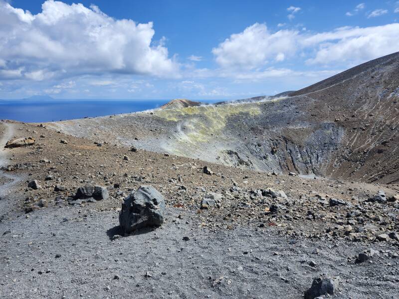 View across a conical crater 500 meters wide and 100 meters deep. On the far side, steam and invisible poisonous gases vent from yellow stone colored by sulfur.