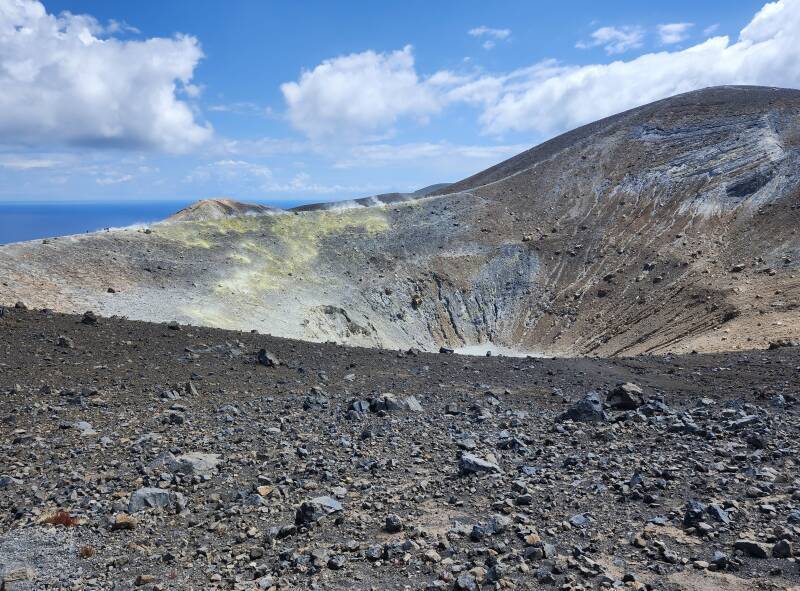 View across a conical crater 500 meters wide and 100 meters deep. On the far side, steam and invisible poisonous gases vent from yellow stone colored by sulfur.