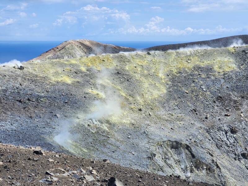 View across a conical crater 500 meters wide and 100 meters deep. On the far side, steam and invisible poisonous gases vent from yellow stone colored by sulfur.