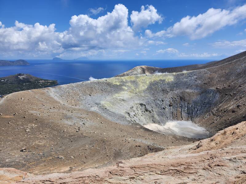View across a conical crater 500 meters wide and 100 meters deep. On the far side, steam and invisible poisonous gases vent from yellow stone colored by sulfur.