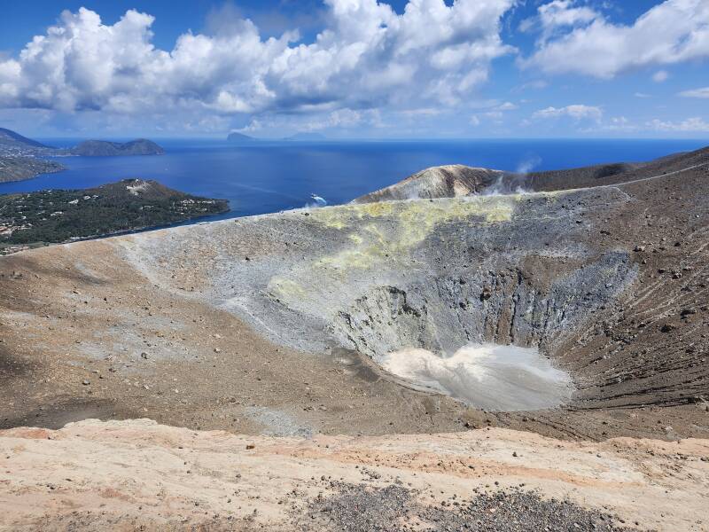 View across a conical crater 500 meters wide and 100 meters deep. On the far side, steam and invisible poisonous gases vent from yellow stone colored by sulfur.