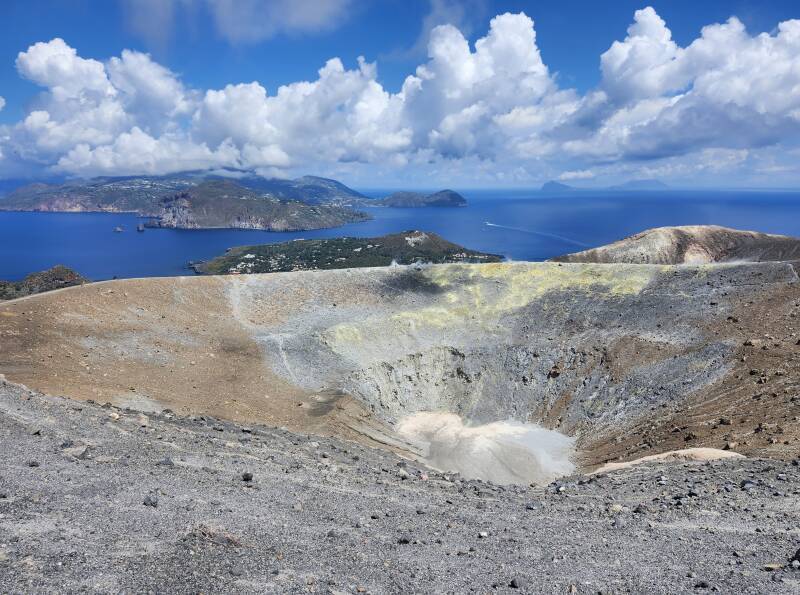 View across a conical crater 500 meters wide and 100 meters deep. On the far side, steam and invisible poisonous gases vent from yellow stone colored by sulfur.