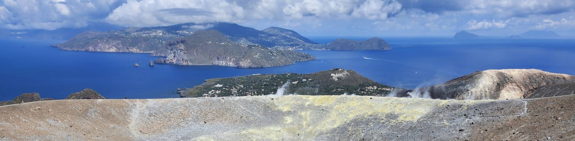 View from the peak of the active volcano on the island of Vulcano: peak crater in foreground, yellow sulfur deposits, small Vulcaneloo crater, open strait of water, then Lipari island, and other islands visible in the distance.