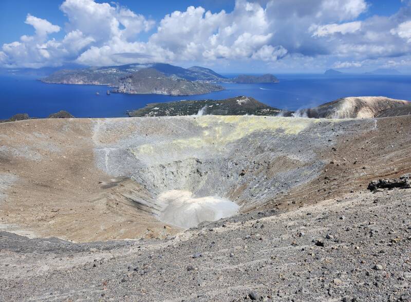 View across a conical crater 500 meters wide and 100 meters deep. On the far side, steam and invisible poisonous gases vent from yellow stone colored by sulfur.