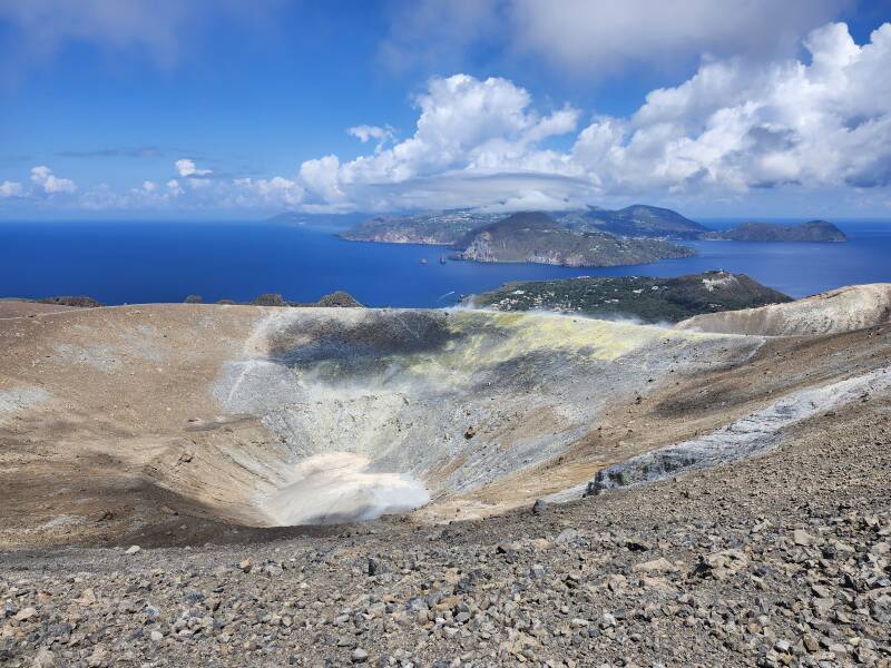 View across a conical crater 500 meters wide and 100 meters deep. On the far side, steam and invisible poisonous gases vent from yellow stone colored by sulfur.