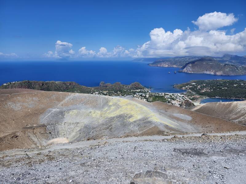 View across a conical crater 500 meters wide and 100 meters deep. On the far side, steam and invisible poisonous gases vent from yellow stone colored by sulfur.
