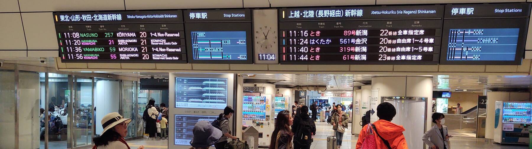 People below an overhead sign showing Shinkansen trains, departure times, and tracks.