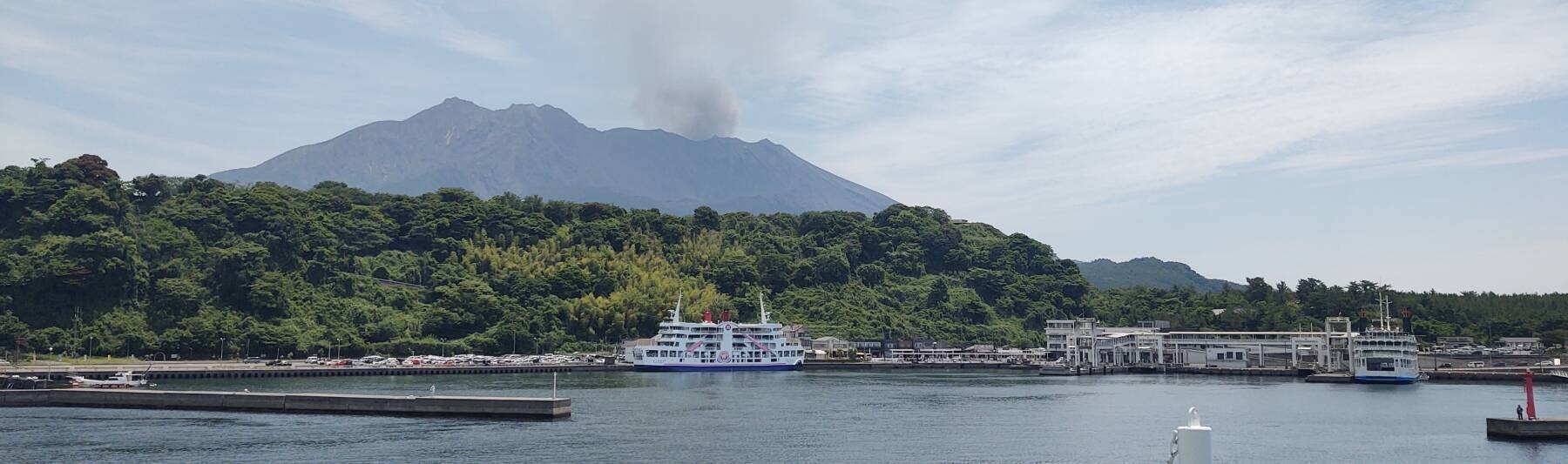 Ferry port with Sakurajima volcano in the distance, an ash plume rising from the southern peak.