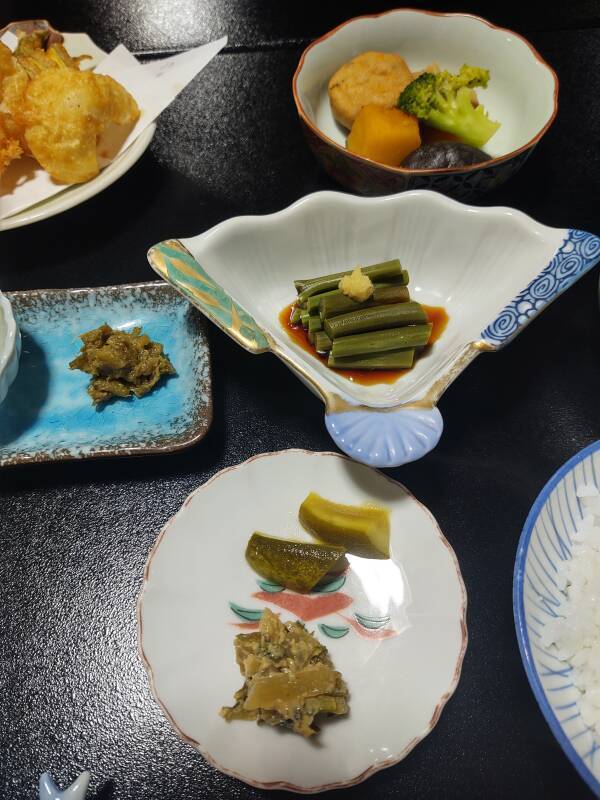 Multiple small dishes of picked vegetables, bamboo shoots, steamed vegetables and mushroom with fish cake.
