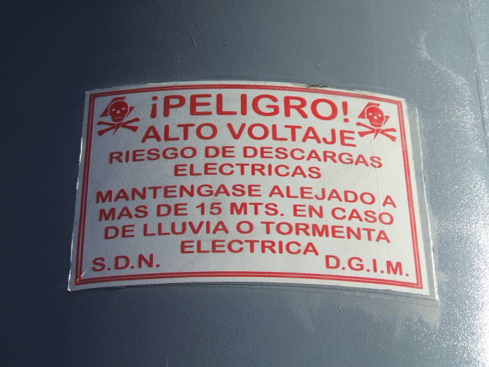 Sign on enormous flag pole: PELIGRO!  ALTO VOLTAJE! Riesgo de descargas electricas.  Mantengase alejado a mas de 15 meters en caso de lluvia o tormenta electrica.