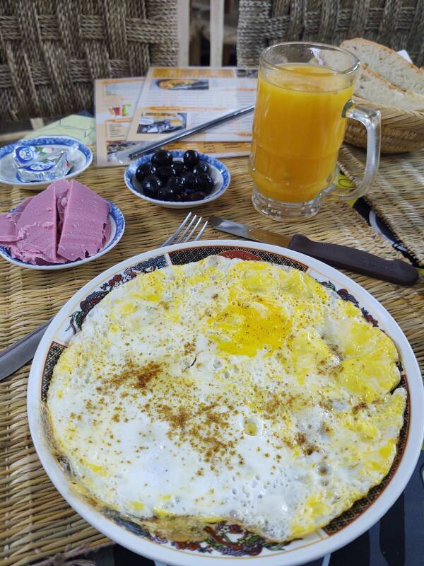 Moroccan breakfast of eggs (omelette), processed meat, olives, cheese, bread, orange juice, and tea.