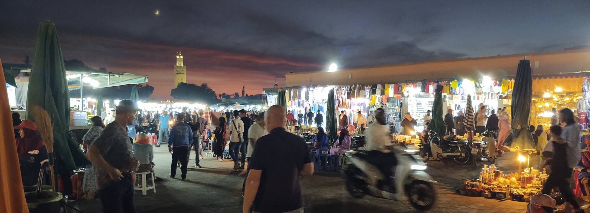 ALT: Vendors, cooks, and visitors on the Jemaa el-Fnaa in Marrakech. In the background, a crescent moon shines above the minaret of the Kutubiyya Mosque.