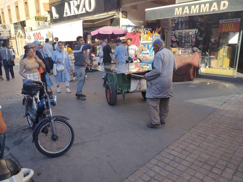 A man prepares to transport a large sheet of glass through a crowded medina as a younger man tells him that his plan will never work.