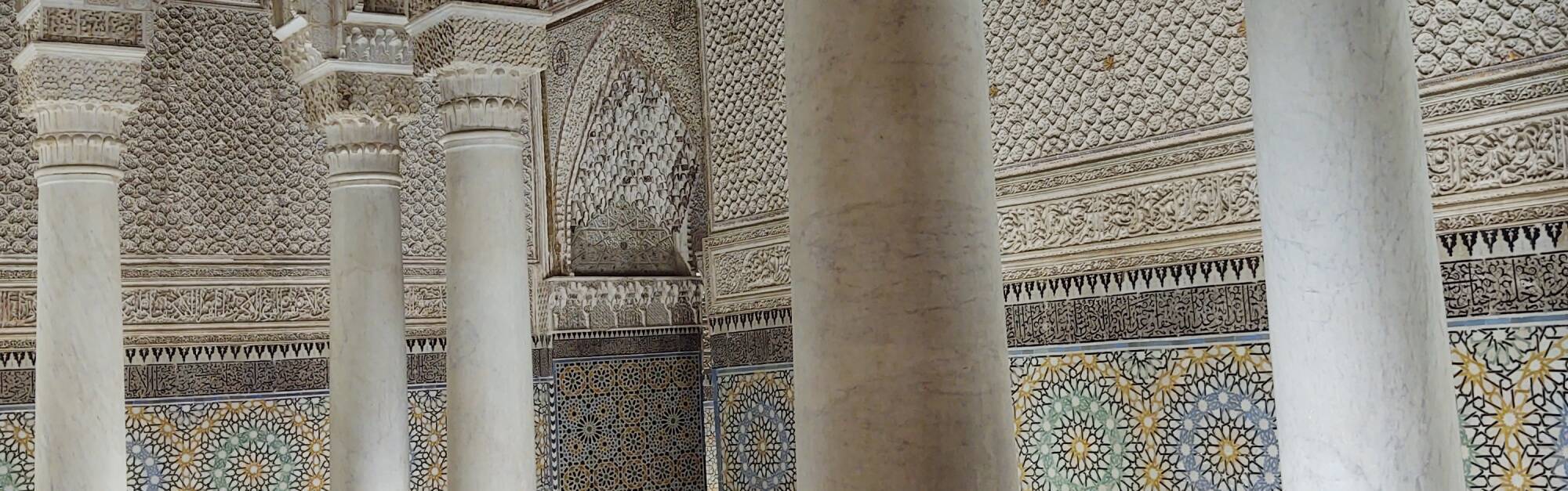 Interior of the Saadian tombs: marble pillars, multi-color zellij tilework, carved stucco. Quranic and devotional text as black on grey tiles, and above that, carved into the stucco. Doorway with lambrequin arch, intricate pattern of lobes and points.