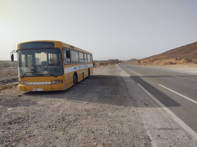 School bus parked along the highway across from a fuel station and rest stop, a ksar or fortified town is in the distance.