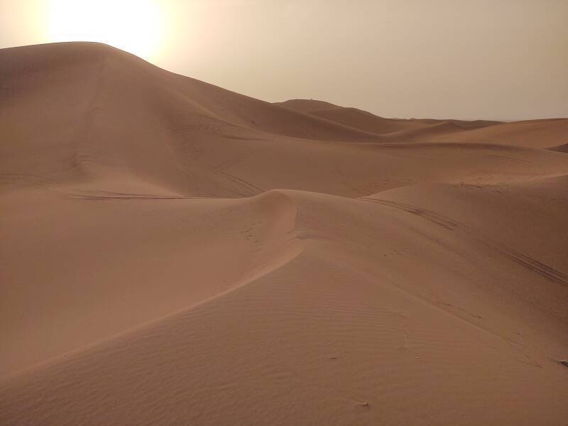 People on top of a 100-meter dune in the distance, off-road vehicle tracks in the foreground, sun low above the dunes.