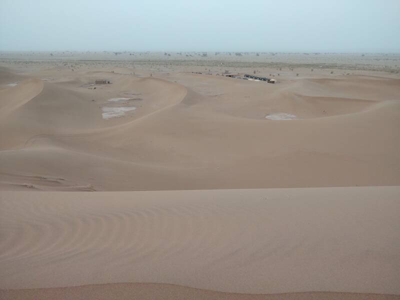 Two camps between low dunes along the north edge of Erg Chigaga. Open bled and acacia trees beyond that.