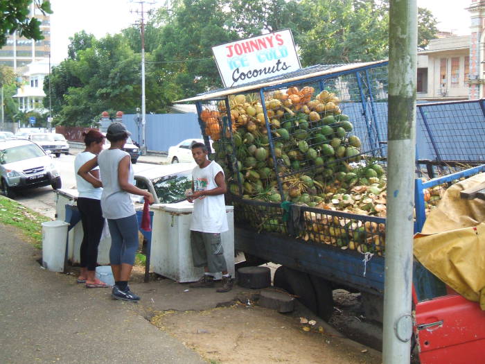 Coconut vendors in Trinidad.  A manu is selling coconuts from a heavily loaded blue truck that is tipped toward the sidewalk.  His sign says 'Johnny's Ice Cold Coconuts'.
