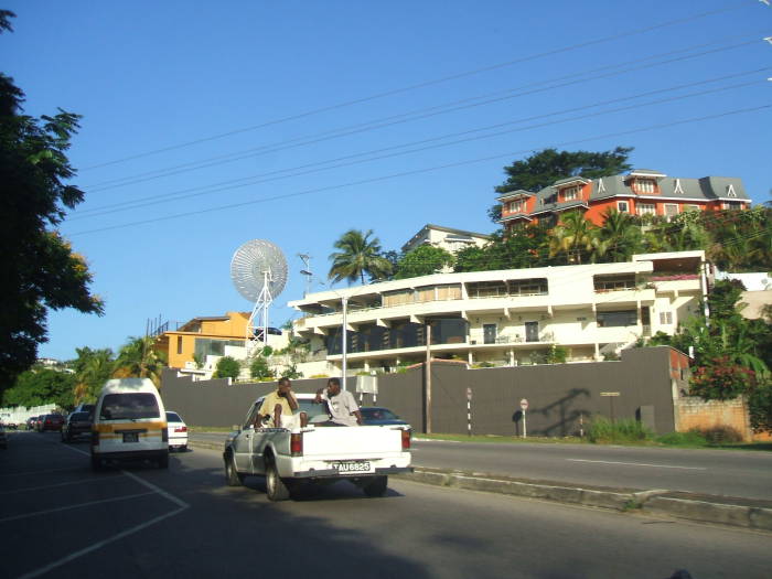 Driving along the highway in Trinidad:  Traffic moves on the left, British style.  A large satellite dish antenna is on a tripod above some apartments.