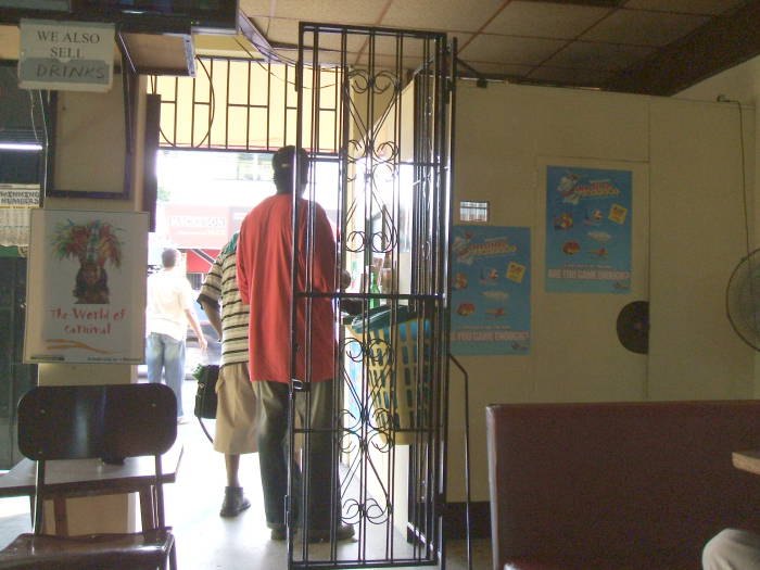 Hanging out with the locals at Bar 52 in western Port of Spain, Trinidad.  Some other patrons talk.  The sign says: 'WE ALSO SERVE DRINKS.'