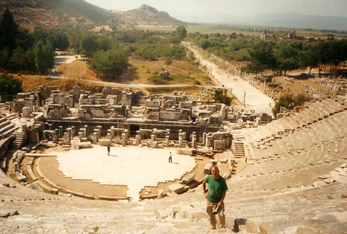 Ephesus: the Great Theatre.  Several rows of marble seats, a marble stage, ruins of the theatre backdrop.  In the distance a road leads past marble columns past the basilica and toward the former harbor district.
