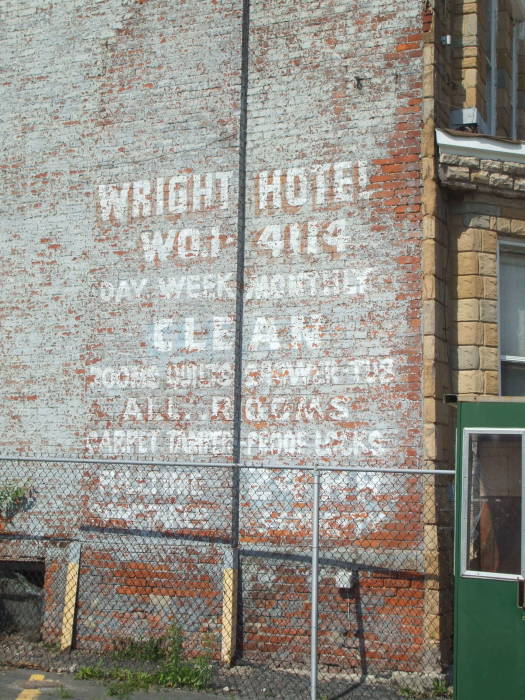 Faded sign painted onto brick:  WRIGHT HOTEL / WO1-4114 / DAY WEEK MONTHLY / CLEAN / ROOMS TOILETS SHOWER TUB / ALL ROOMS / CARPET TAMPER-PROOF LOCKS / FULL TIME 24 HR / CARETAKER SECURITY.
