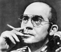 Hunter S Thompson, smoking a cigarette in a holder and wearing tinted aviator glasses.